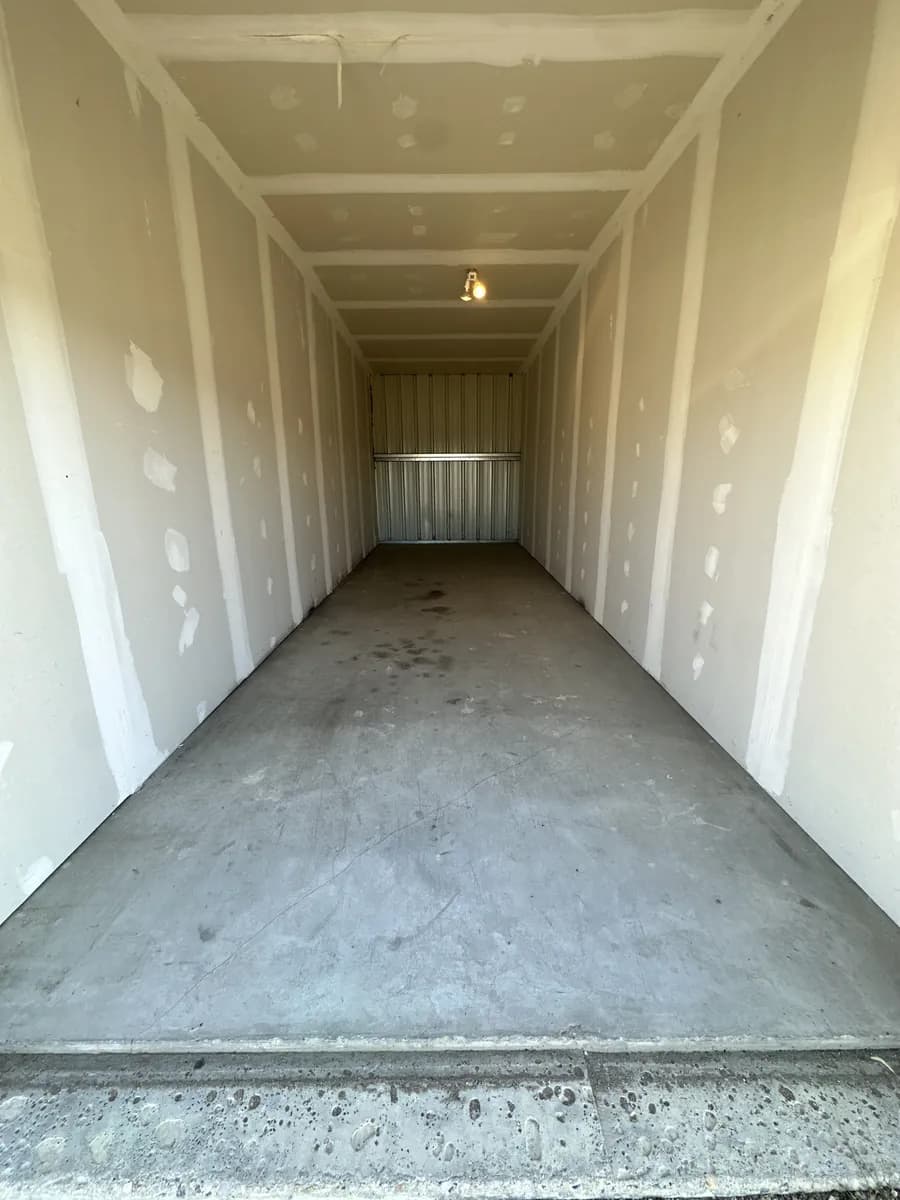 Inside a long, empty storage bay with drywall walls, a ceiling light and a concrete floor