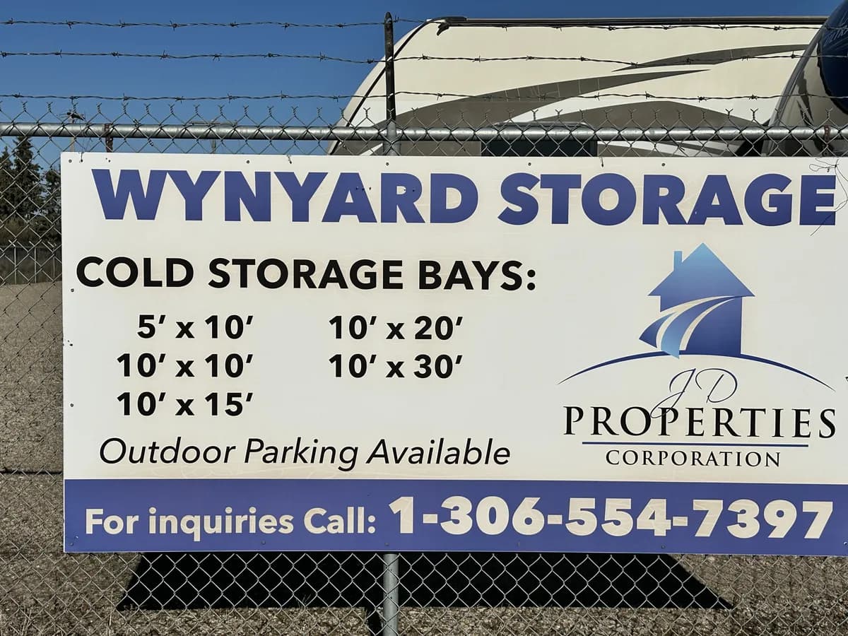 Wynyard Storage sign on the fence: cold storage bays 5′×10′, 10′×10′, 10′×15′, 10′×20′ and 10′×30′, outdoor parking available, JD Properties Corporation logo, and 'For inquiries call 1-306-554-7397'